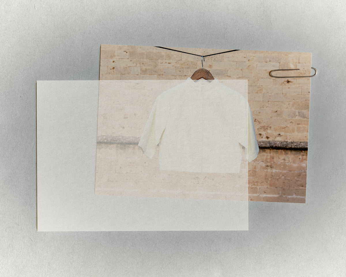 A cream shirt on a wooden hanger against a stone wall, the photograph clipped to a sheet of paper with a sheet of glassine laid over it